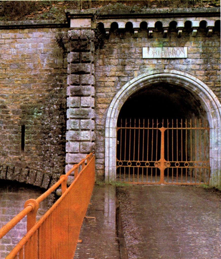 L'entrée du fort de Bron fermé par une grille de couleur orange. Au premier plan le pont dormant en béton permet de traverser le fossé.